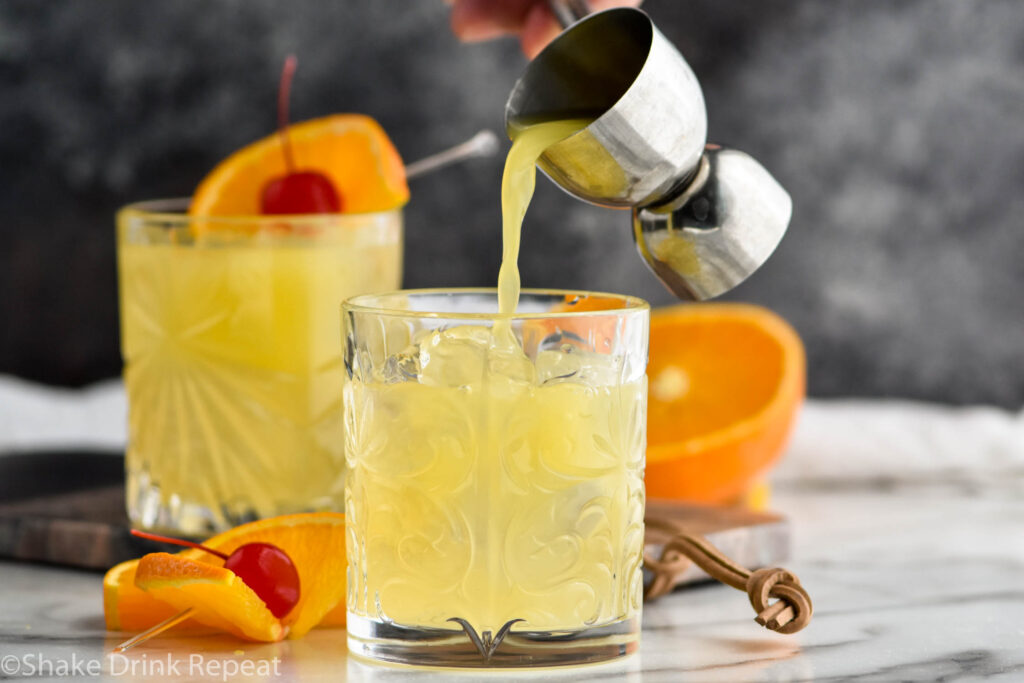 two glasses of Amaretto Stone Sour with jigger of orange juice pouring into glass with orange slice and cherry garnish