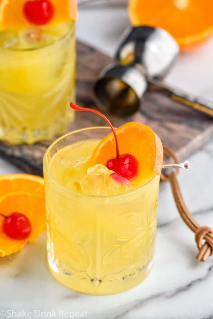 two glasses of Amaretto Stone Sour with ice and fresh orange slice and cherry garnish with jigger and orange in the background