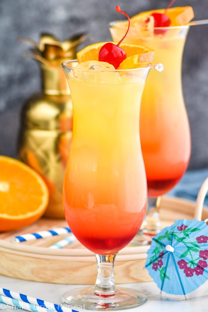two glasses of bahama mama with ice, fresh orange slices, and cherry garnish surrounded by drink umbrella and straws