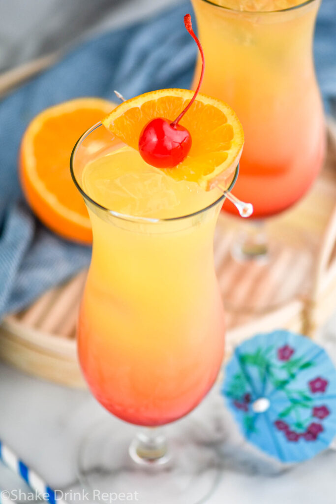 glass of bahama mama with ice, orange slice, and cherry garnish surrounded by drink umbrella and straws