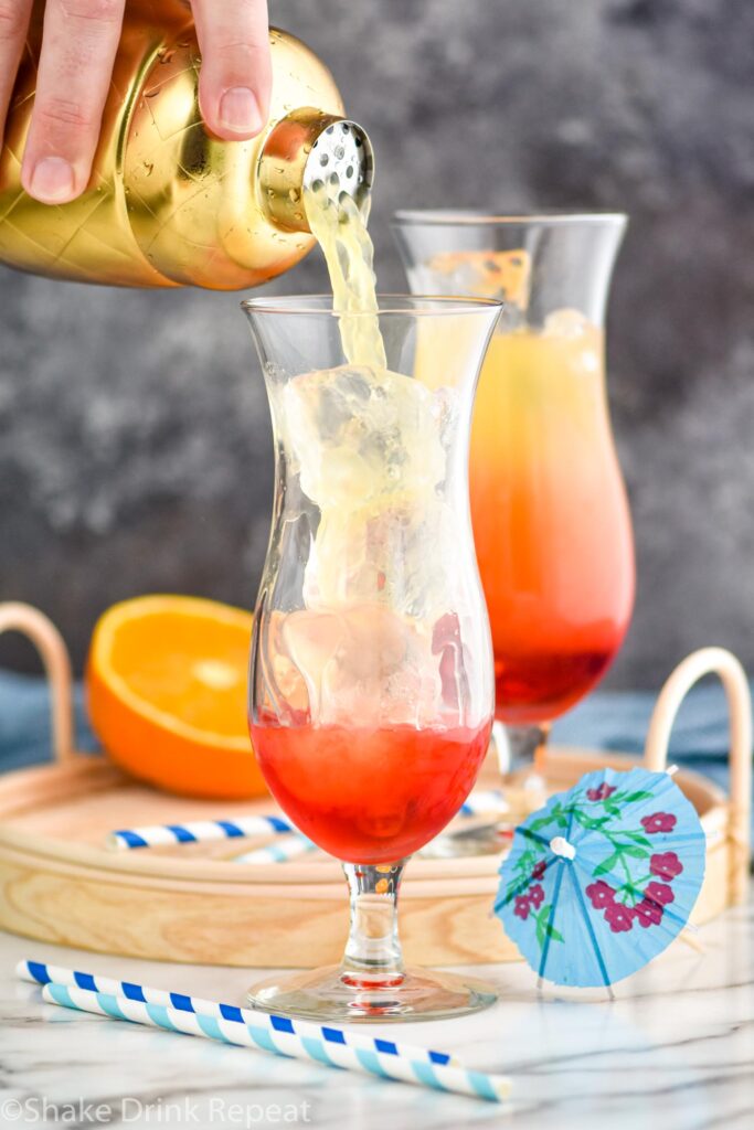 man pouring bahama mama ingredients from a shaker into a glass filled with ice surrounded by straws and a cocktail umbrella