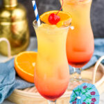 two glasses of bahama mama with ice, straws, fresh orange slices and cherries