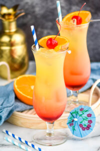 two glasses of bahama mama with ice, straws, fresh orange slices and cherries