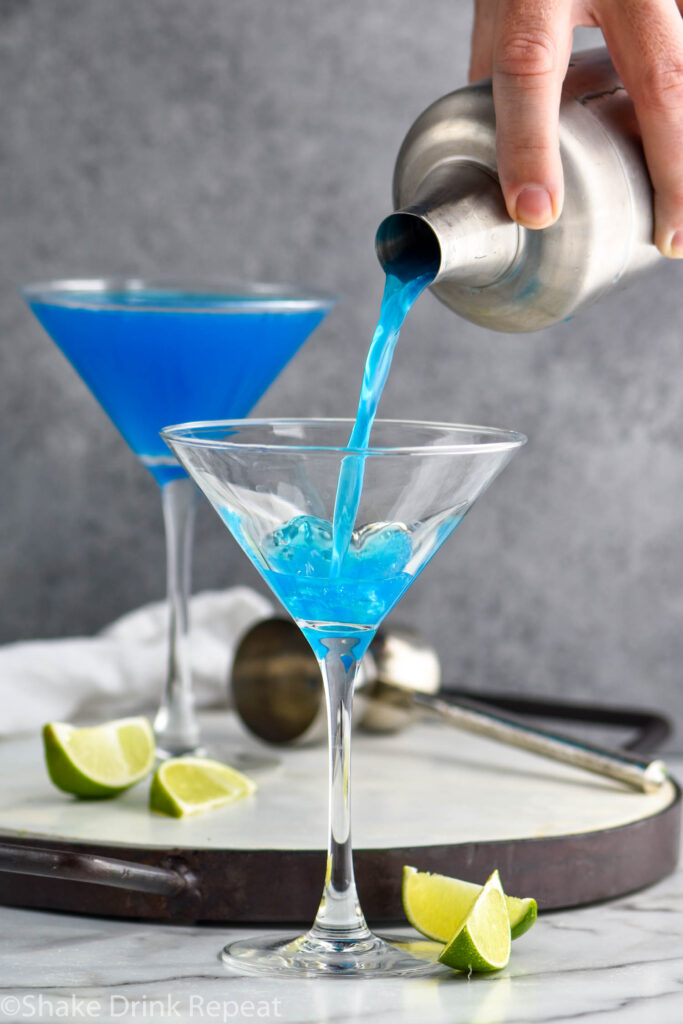 man pouring shaker of blue daiquiri ingredients into a martini glass surrounded by fresh lime wedges and a jigger