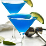 two martini glasses of blue daiquiri with fresh lime wedge garnish and jigger in background