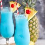 two glasses of blue hawaiian with ice, straws, fresh pineapple, and cherries
