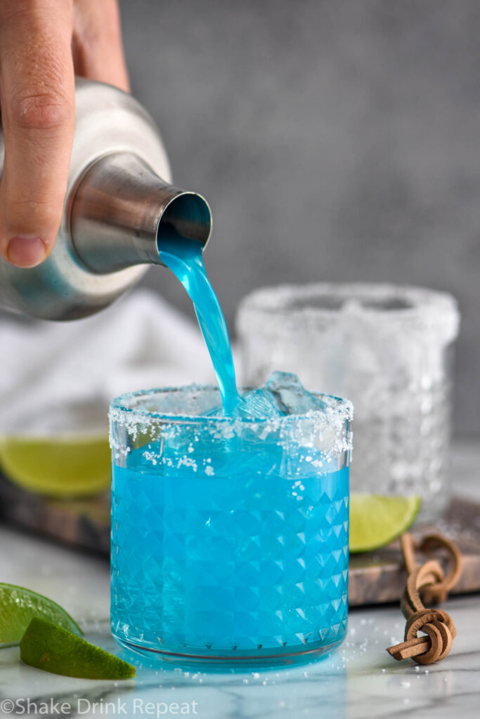 man pouring blue margarita from a shaker into a glass with ice, salted rim and fresh lime wedges