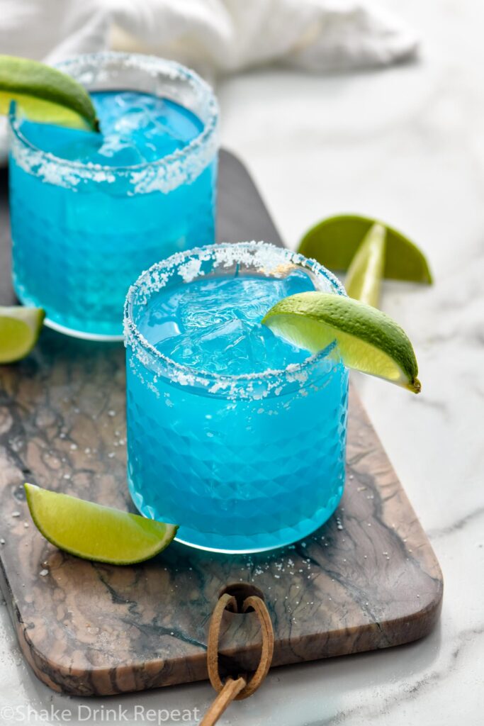 two glasses of blue margarita with salted rim, ice, and fresh lime wedges