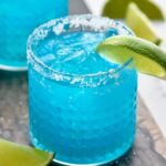 two glasses of blue margarita with ice, salted rim, and fresh lime wedge