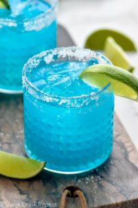 two glasses of blue margarita with ice, salted rim, and fresh lime wedge