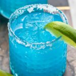 two glasses of blue margarita with salted rims, ice, and fresh lime wedge