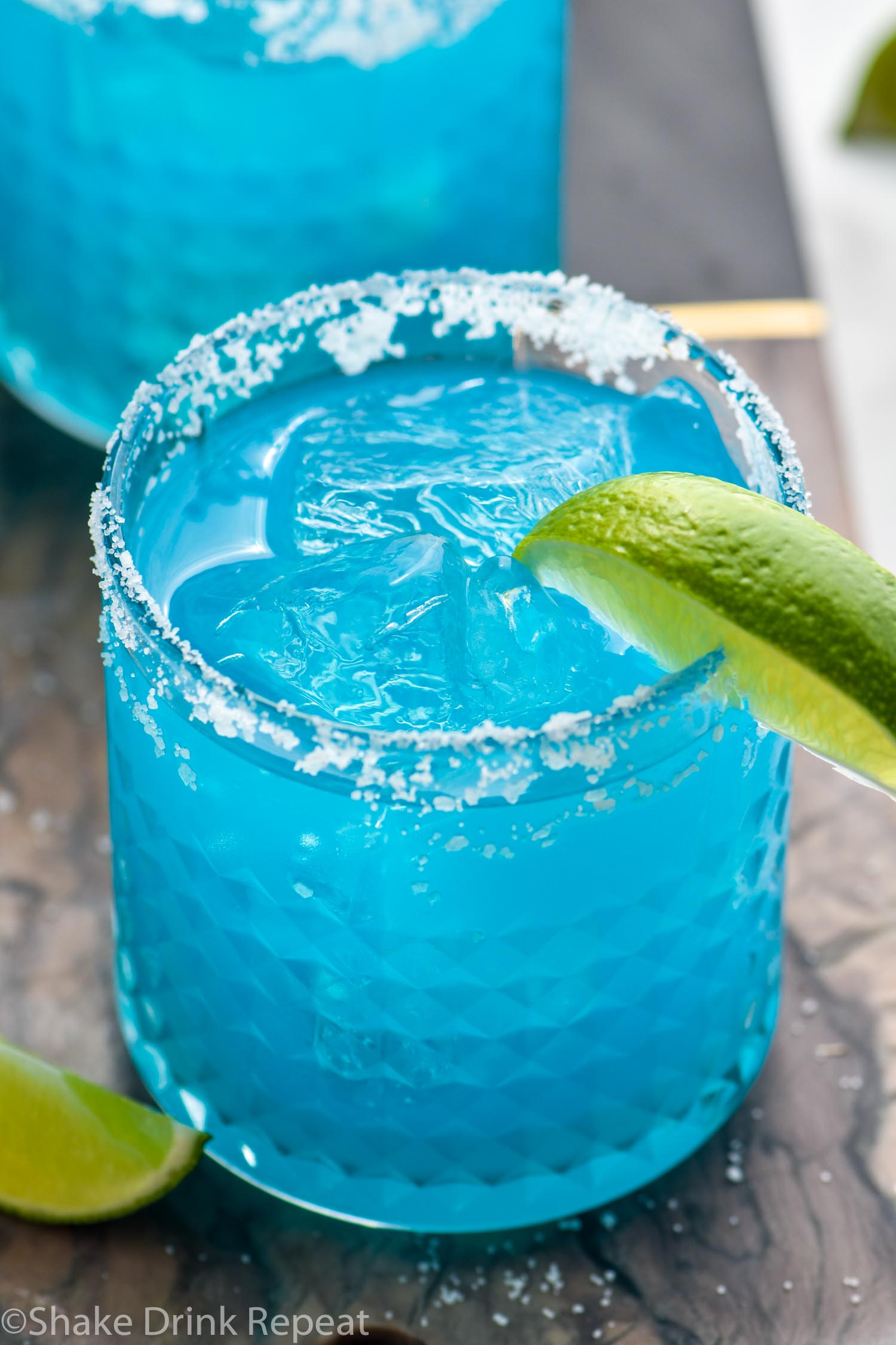 two glasses of blue margarita with salted rims, ice, and fresh lime wedge