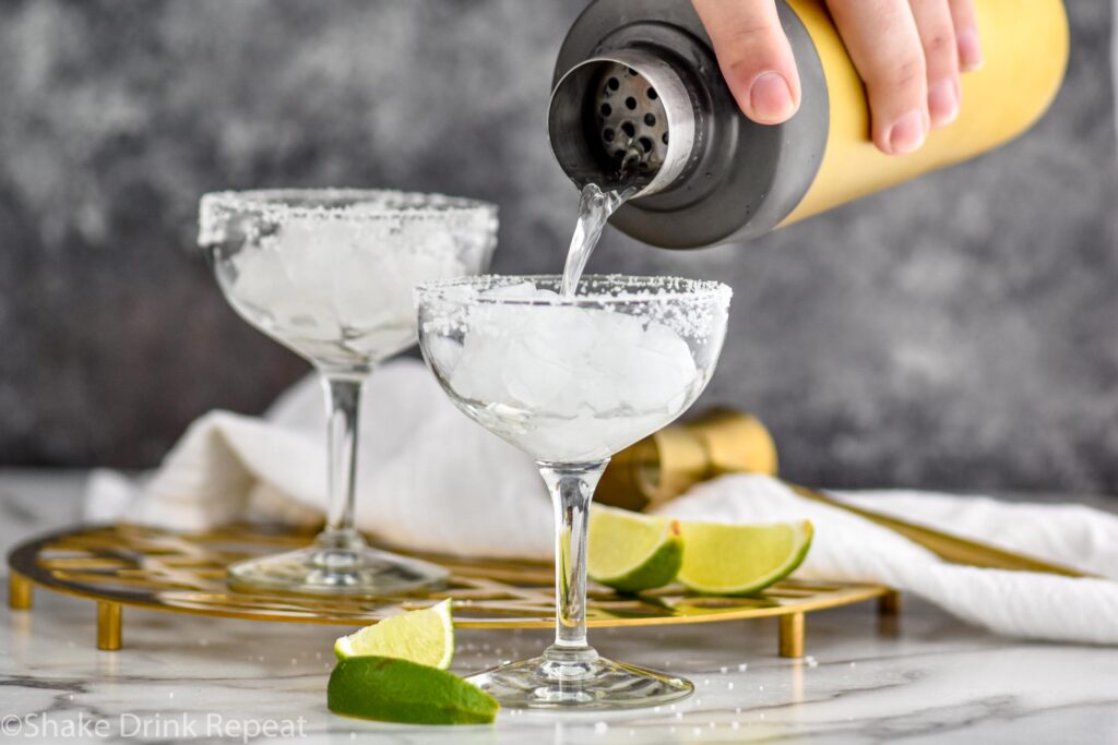 Man's hand pouring shaker of Cadillac Margarita ingredients into a margarita glass with salted rim filled with ice and surrounded by lime wedges