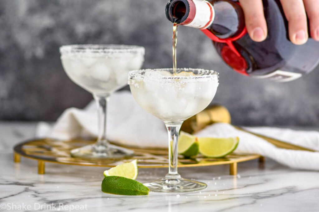 man pouring Grand Marnier into a glass of cadillac margarita with ice, salted rim, and lime wedges