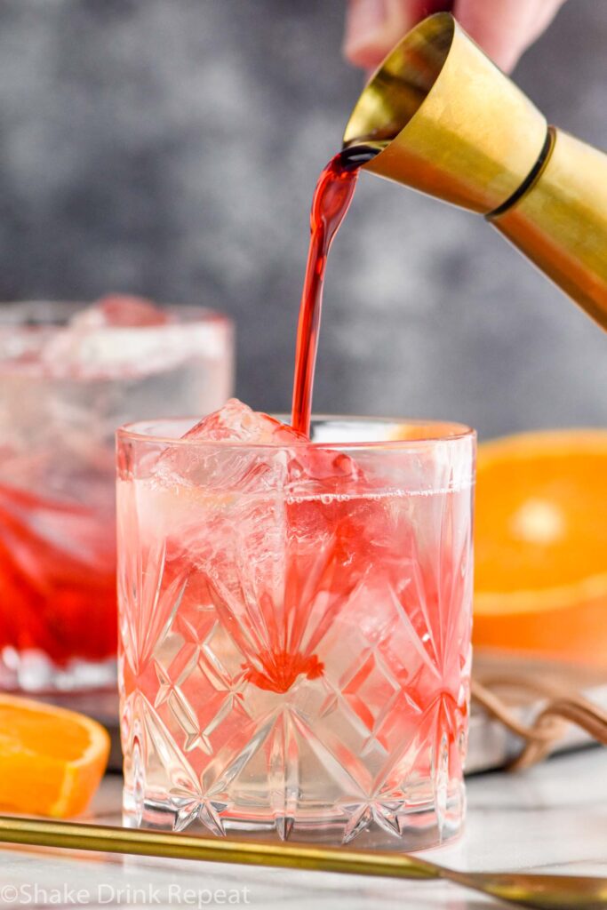 man's hand pouring jigger of grenadine into a glass of cherry vodka soda and ice surrounded by a spoon and fresh orange slices