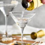 man's hand pouring shaker of Gibson ingredients into a martini glass surrounded by cocktail onions