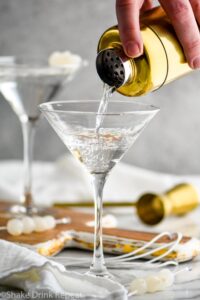 man's hand pouring shaker of Gibson ingredients into a martini glass surrounded by cocktail onions