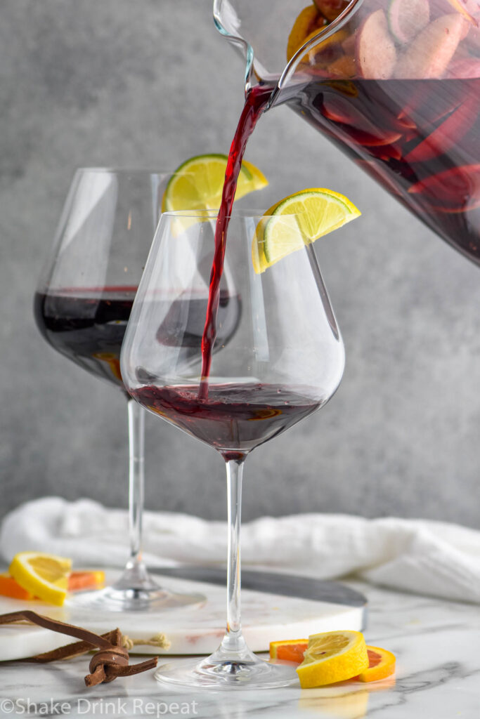 pouring a pitcher of red sangria into two wine glasses with slices of fresh fruit including oranges, lemons, and limes