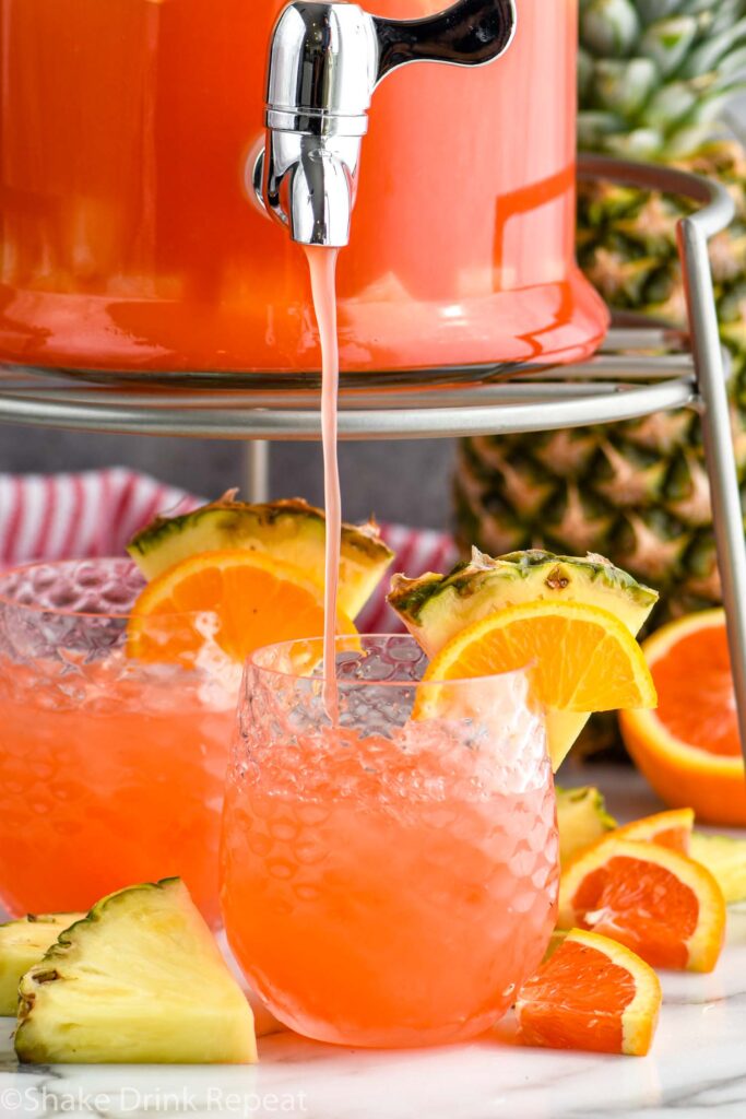 Glass dispenser of of rum punch pouring into a glass of ice with fresh pineapple and orange slices