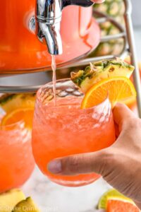 man holding a glass of rum punch with ice and fresh pineapple and orange slice while it fills from dispenser.