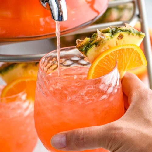 man holding a glass of rum punch with ice and fresh pineapple and orange slice while it fills from dispenser.