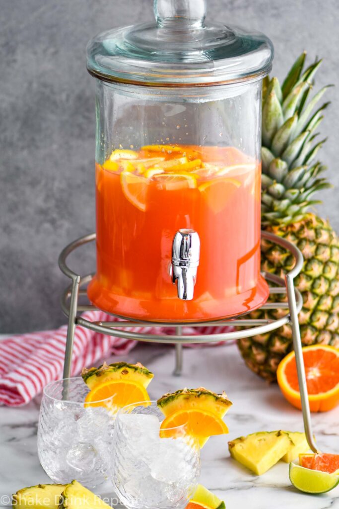 glass dispenser of rum punch with two glasses of ice and slices of oranges, limes, and pineapple