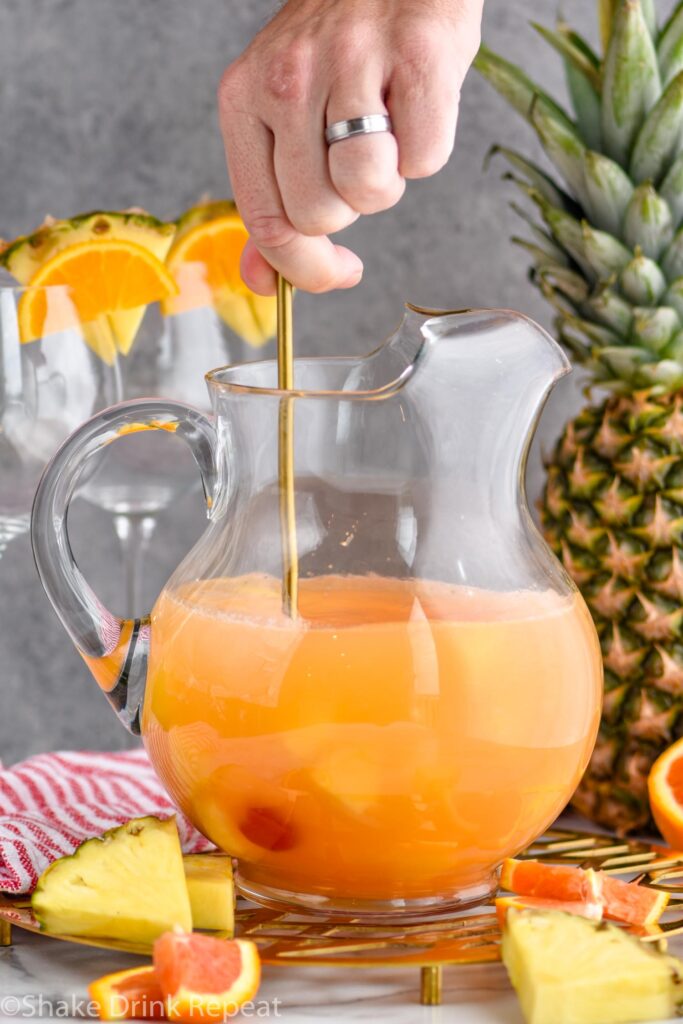 man's hand stirring pitcher of tropical sangria surrounded by slices of fresh pineapple and oranges