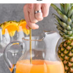 man's hand stirring pitcher of tropical sangria surrounded by slices of fresh pineapple and oranges