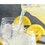 pouring lemonade into a glass of ice to make a vodka lemonade with lemon garnish