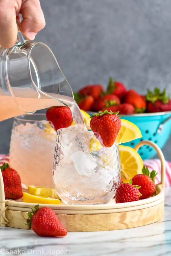 man pouring a pitcher of vodka strawberry lemonade into a glass of ice with fresh strawberries and lemon slices