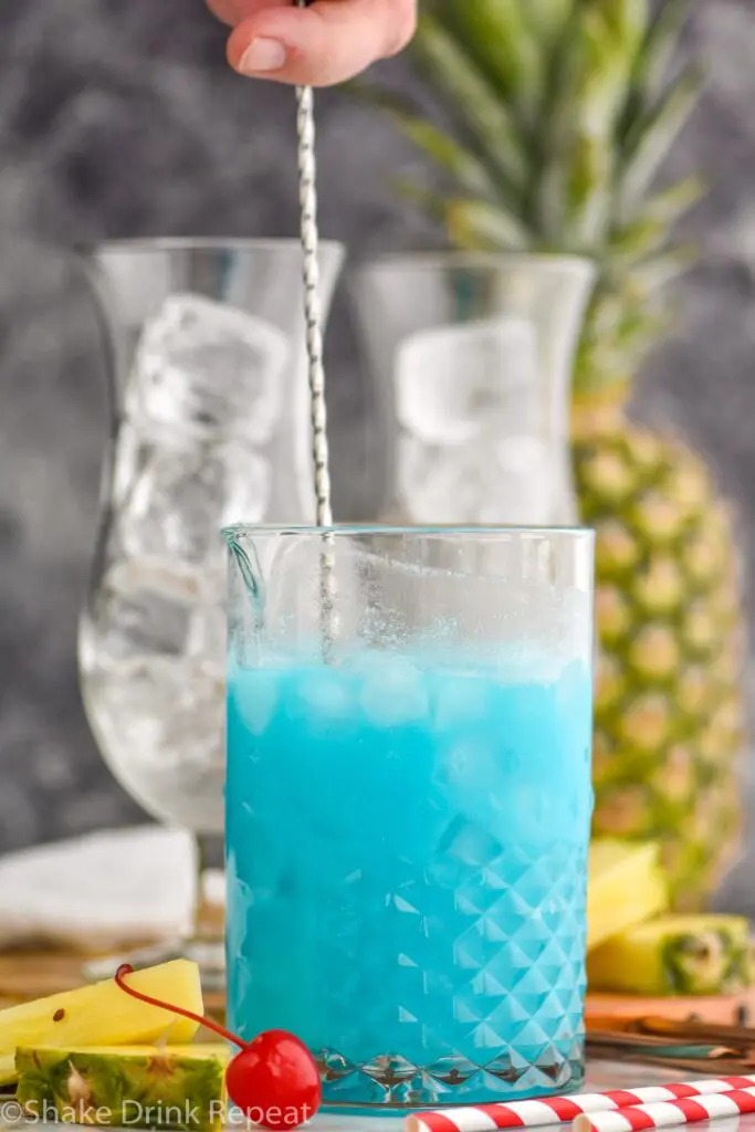 man stirring ingredients of blue hawaiian in a glass with ice surrounded by straws, fresh pineapple and cherries