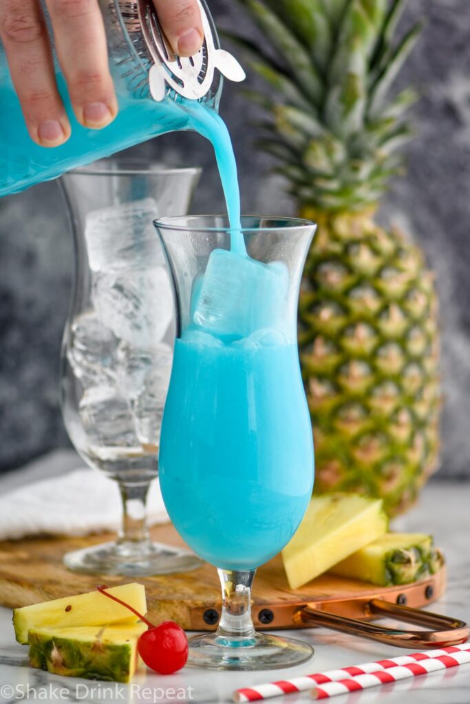 man pouring blue hawaiian ingredients from a shaker into a glass filled with ice surrounded by straws, fresh pineapple wedges and cherries