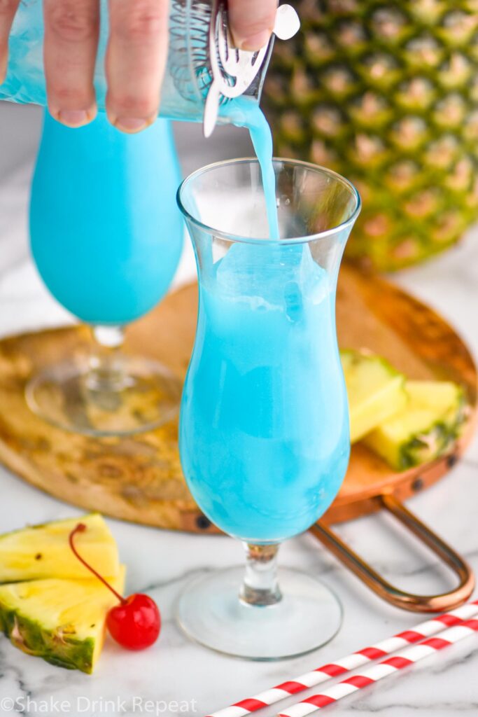 man pouring blue hawaiian ingredients from a shaker into a glass filled with ice surrounded by straws, fresh pineapple wedges and cherries.