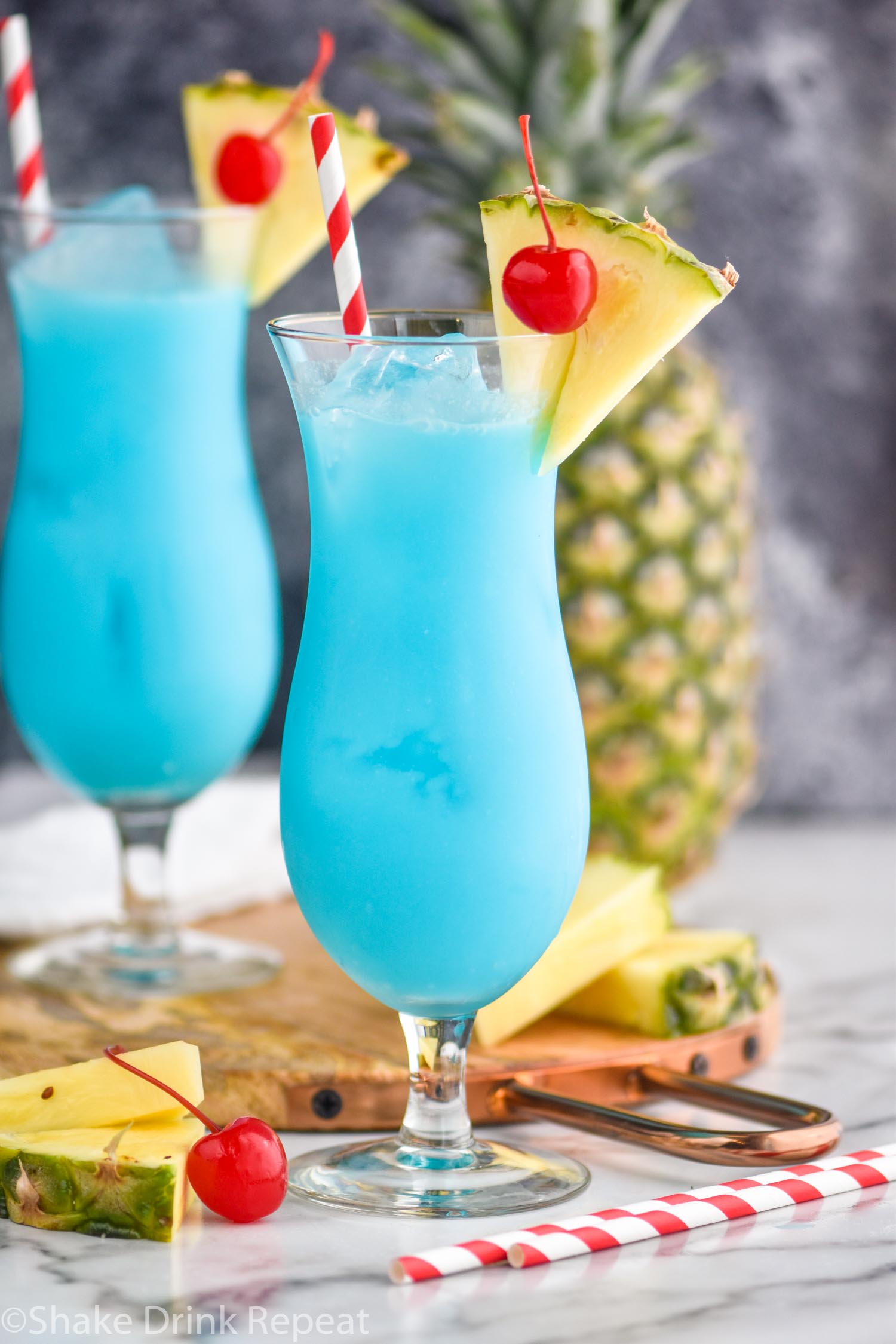 two glasses of blue hawaiian with ice, straws, cherries, and fresh pineapple wedge garnish