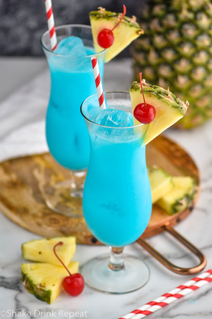 two glasses of blue hawaiian with ice, straws, cherries, and wedges of fresh pineapple