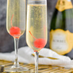 two glasses of french 76 garnished with a cherry with bottle of champagne, cork, and spoon in the background