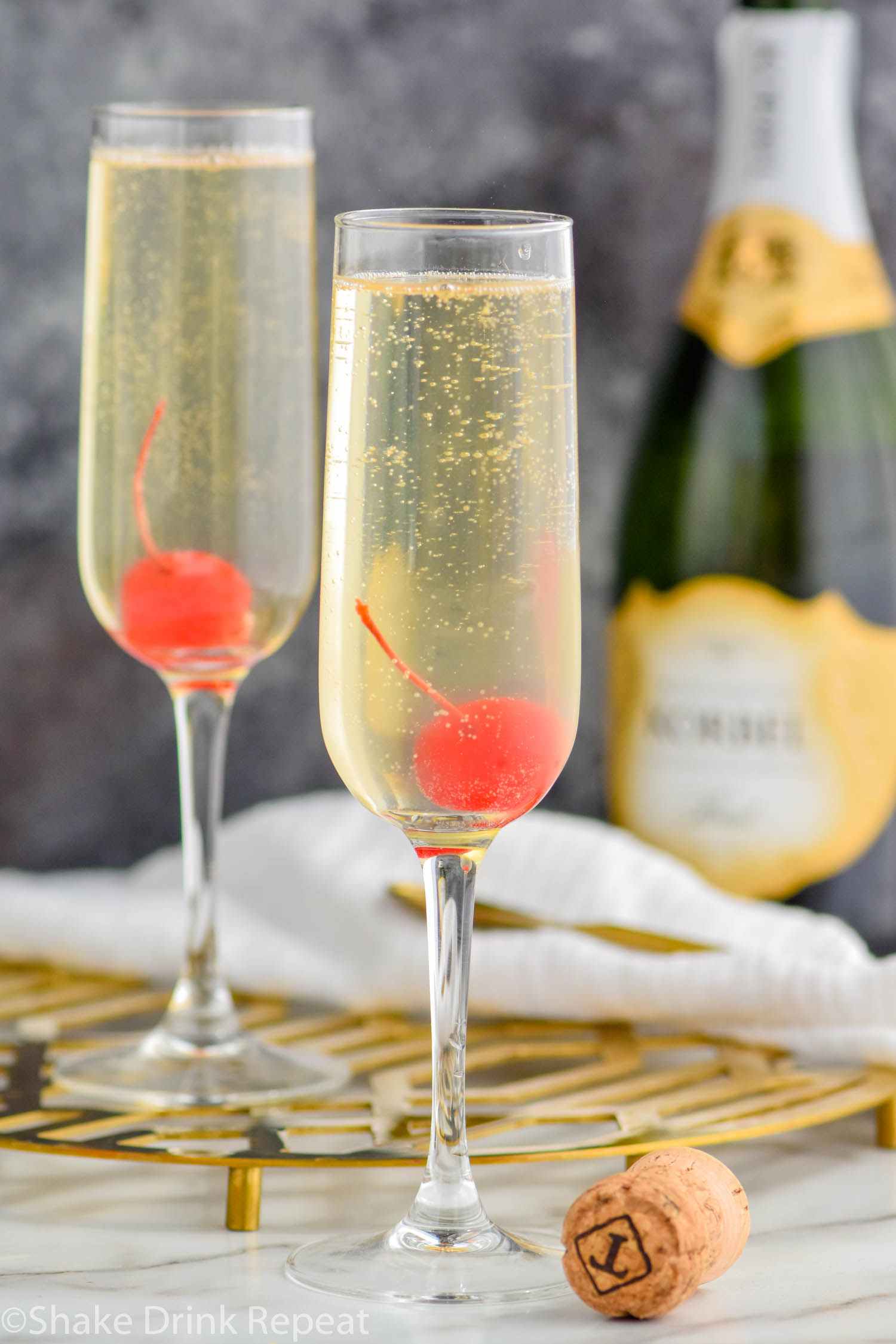 two glasses of french 76 garnished with a cherry with bottle of champagne, cork, and spoon in the background