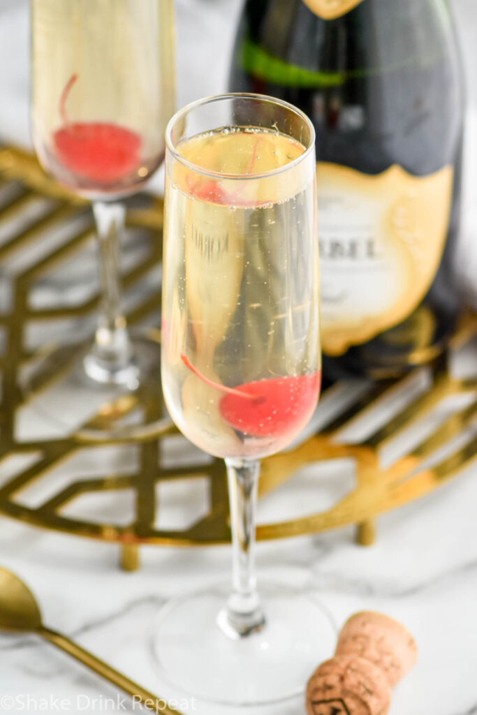 two glasses of french 76 garnished with a cherry surrounded by bottle of champagne, spoon, and cork