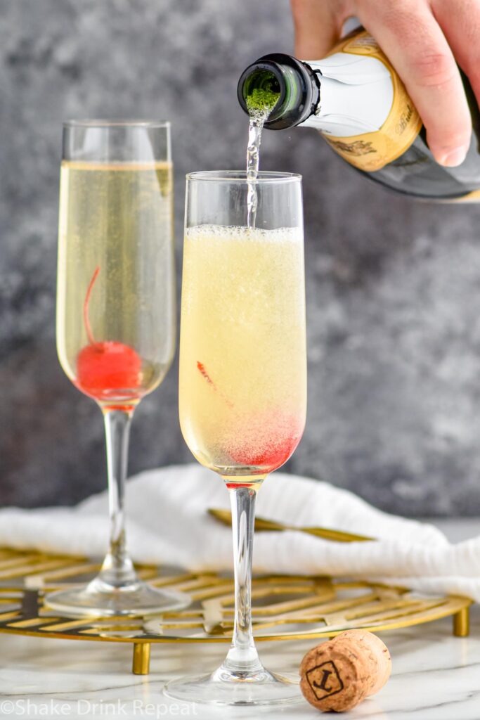 man's hand pouring champagne from a bottle into a glass of french 76 ingredients garnished with a cherry