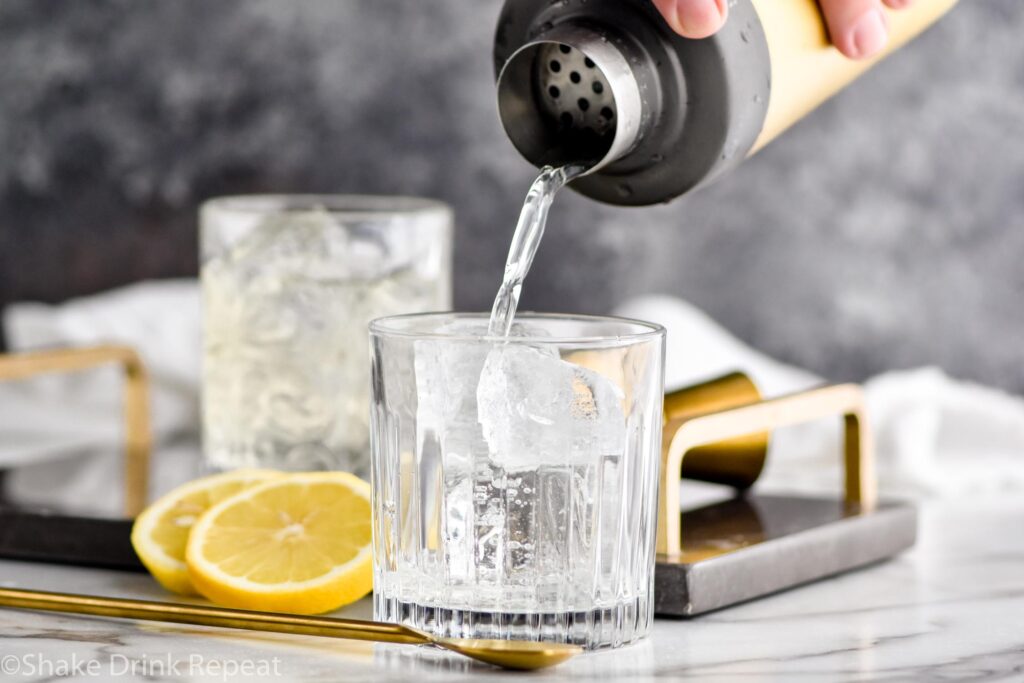 man pouring shaker of vodka sour ingredients into a rocks glass filled with ice surrounded by fresh lemon slices and a spoon