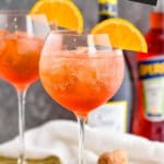 two glasses of Aperol Spritz with ice and fresh orange slices surrounded by two straws, a bottle of prosecco and a bottle of Aperol