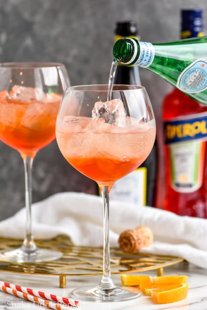 bottle of club soda pouring into a glass of aperol spritz with ice and orange slices surrounded by straws, a bottle of sparkling wine, and a bottle of aperol