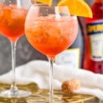 two glasses of aperol spritz with ice and orange slices surrounded by straws, a bottle of sparkling wine, and a bottle of aperol