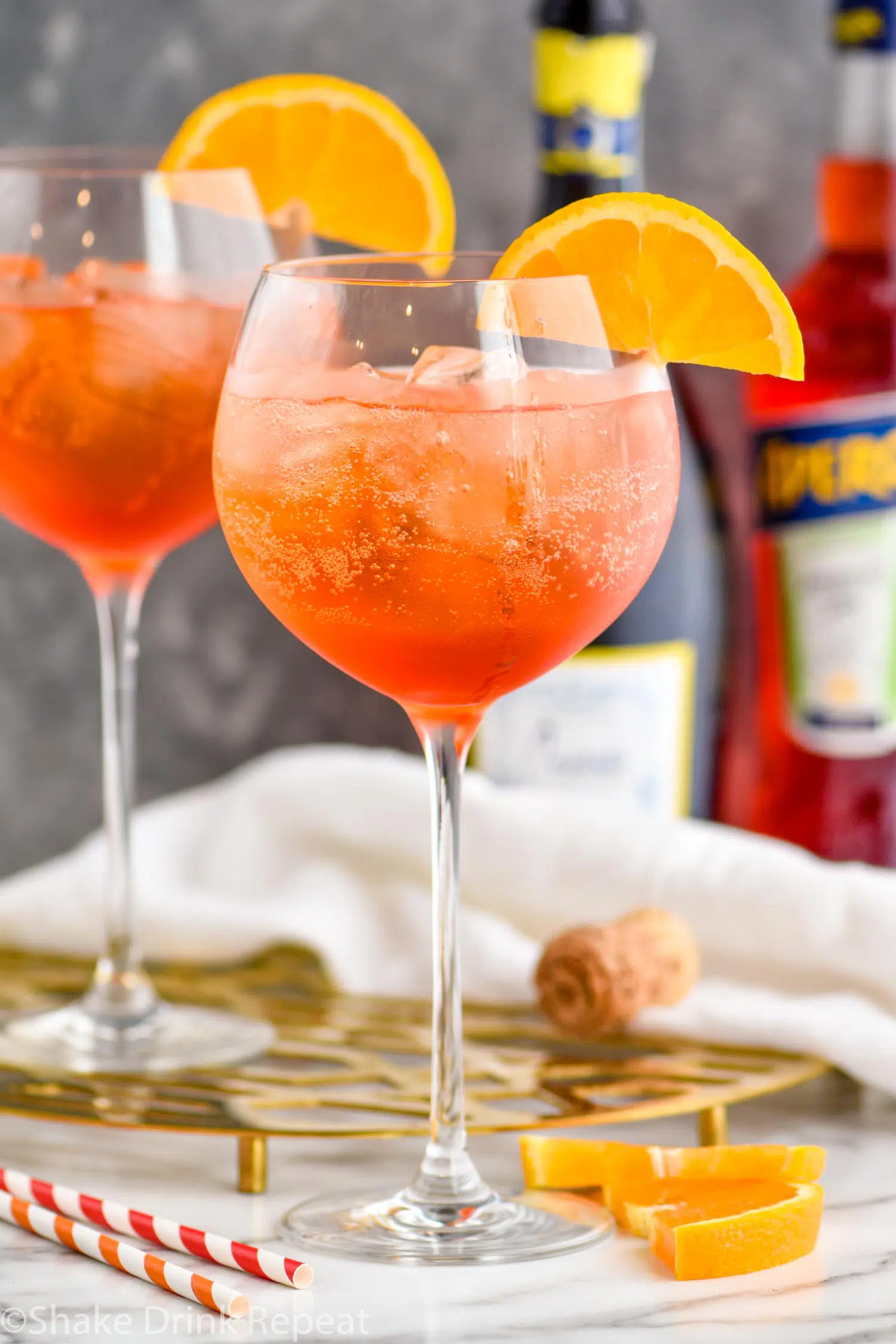 two glasses of aperol spritz with ice and orange slices surrounded by straws, a bottle of sparkling wine, and a bottle of aperol