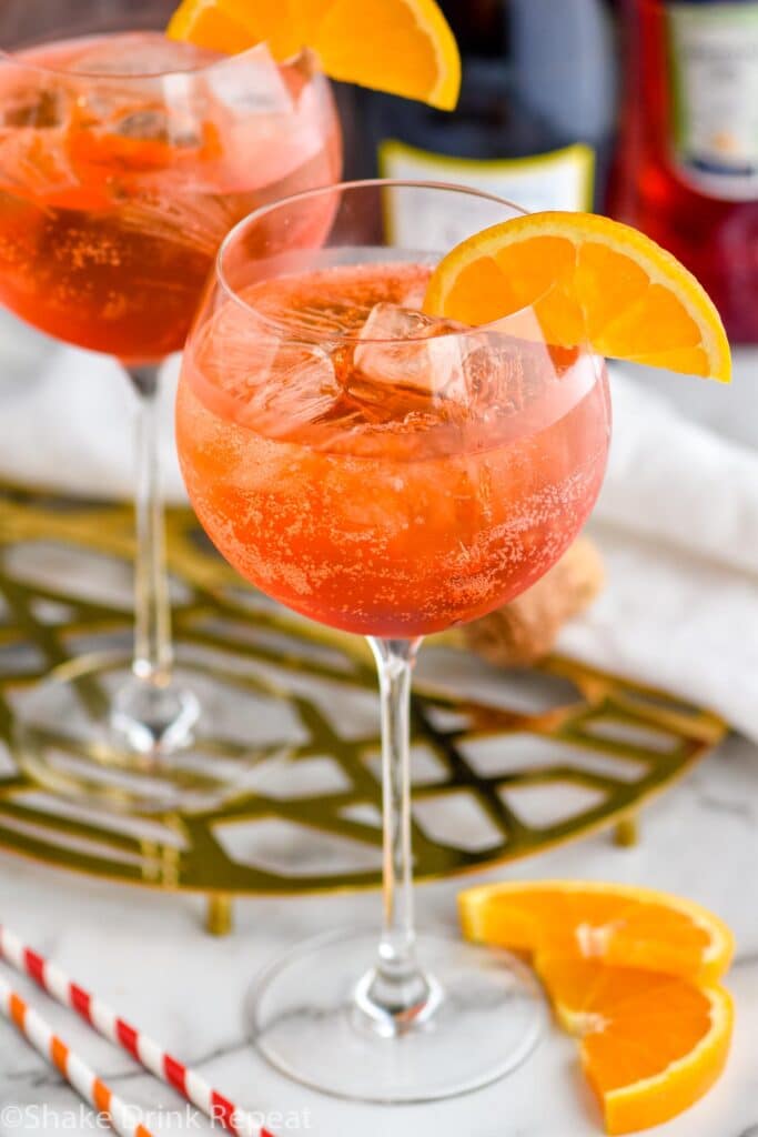 two glasses of aperol spritz with ice and fresh orange slice garnish surrounded by straws and bottle of sparkling wine