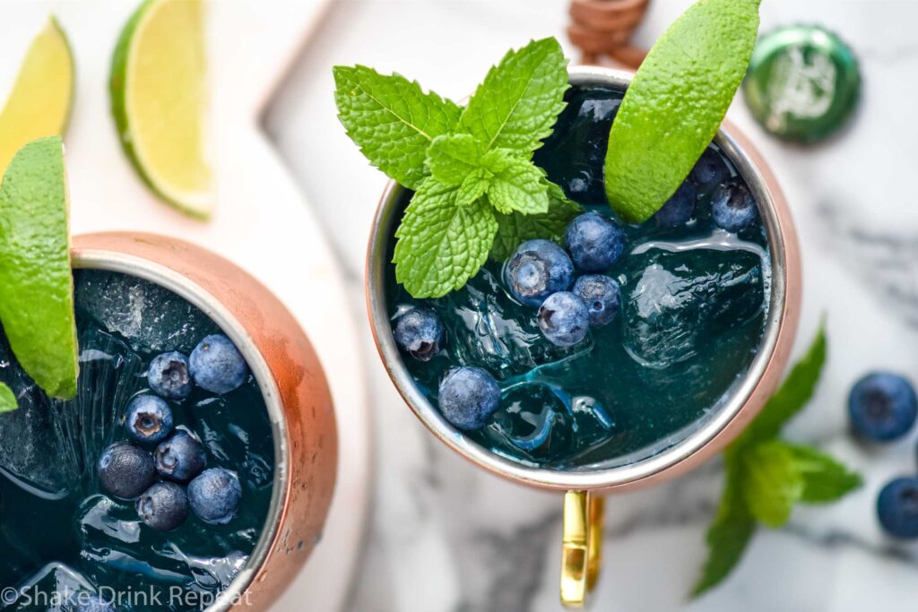 two copper mugs of blue mules with ice, blueberries, lime wedge, and mint leaves