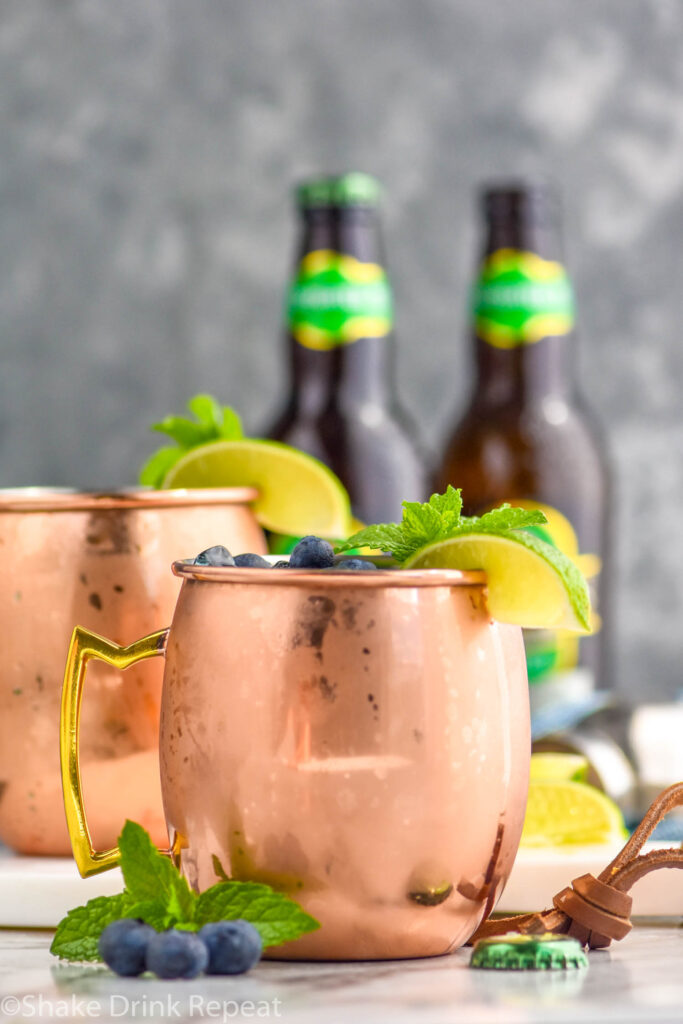 two copper mugs of blue mules with ice, blueberries, lime wedge, and mint leaves and two bottles of ginger beer in the background