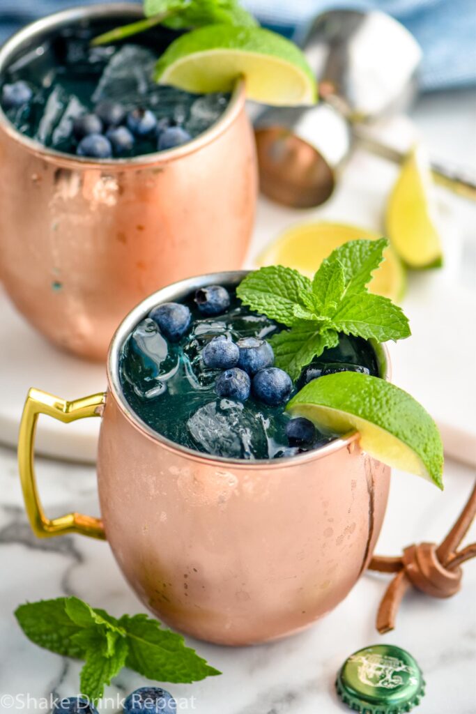 two copper mugs of blue mules with ice, blueberries, lime wedge, and mint leaves