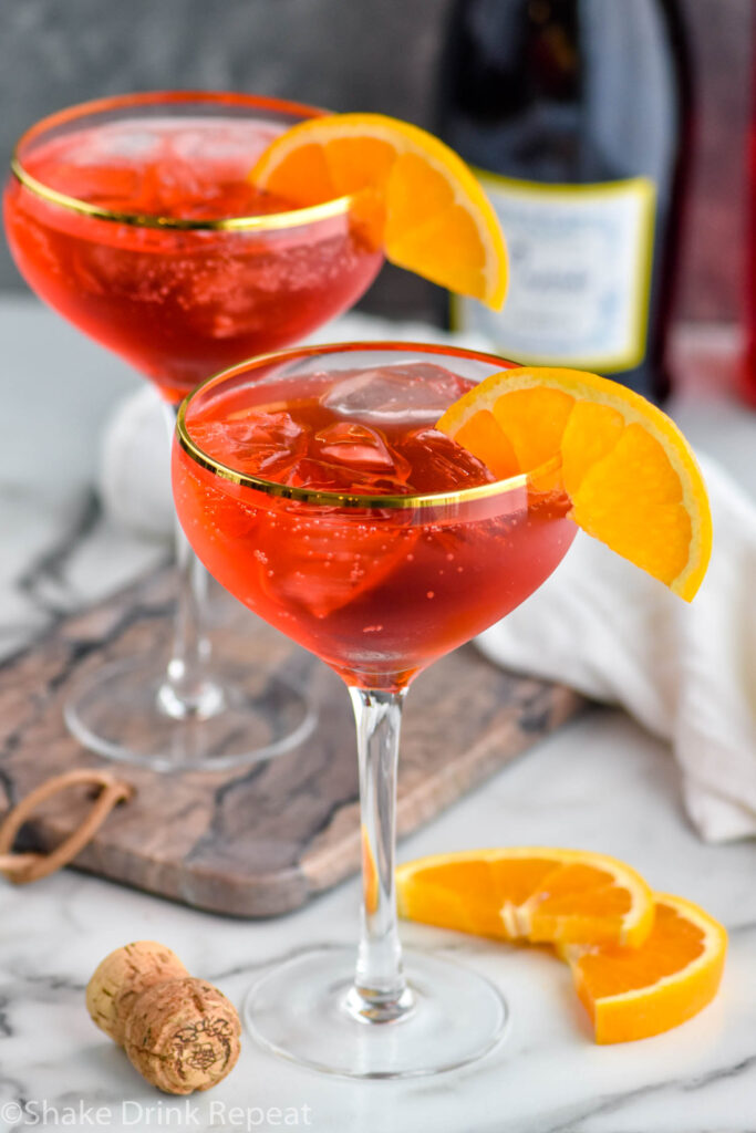 two glasses of Campari Spritz with ice and fresh orange slices surrounded by cork and bottle of sparkling wine