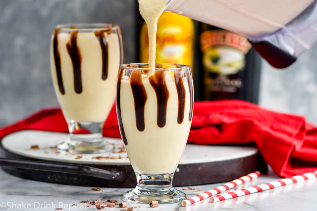 blender pouring Frozen Mudslide recipe into a glass drizzled with chocolate syrup surrounded by chocolate shavings, straws, bottle of Baileys and bottle of Kahlua